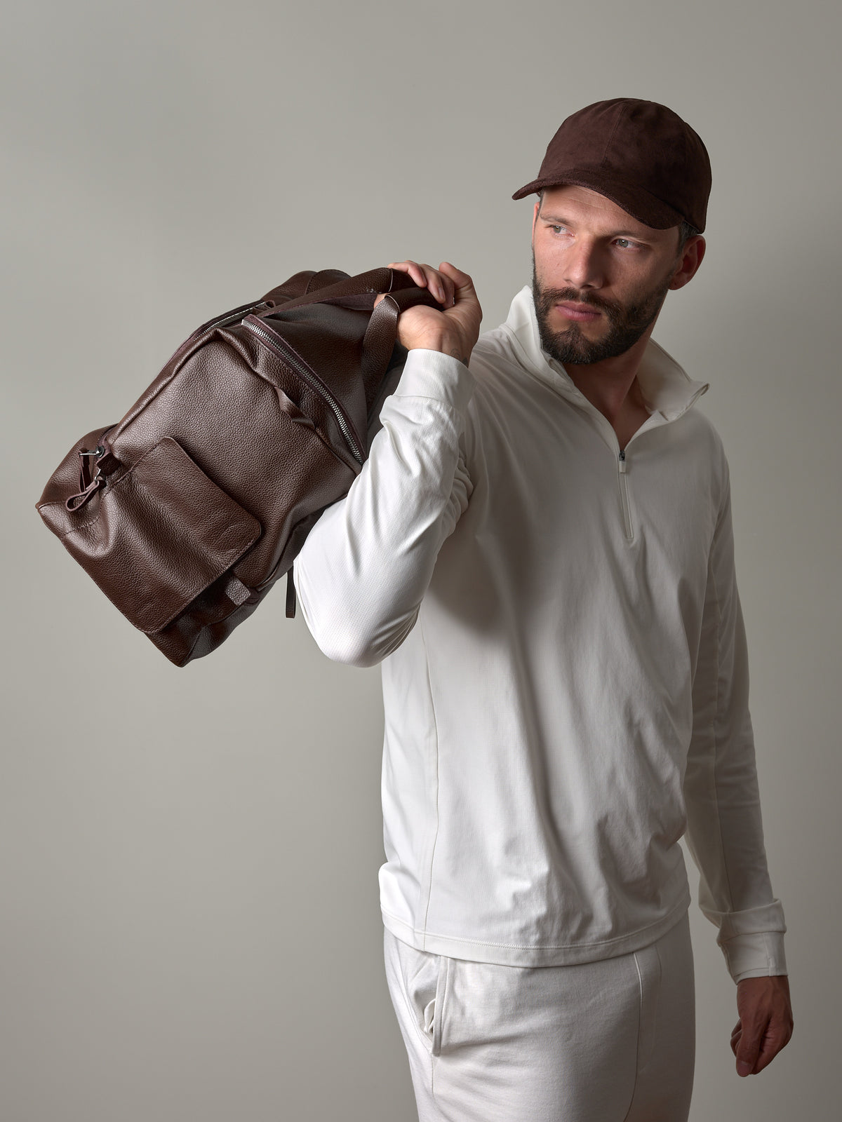 A man wearing a white shirt and brown cap carrying a dark brown Capra Ventra Holdall over his shoulder.