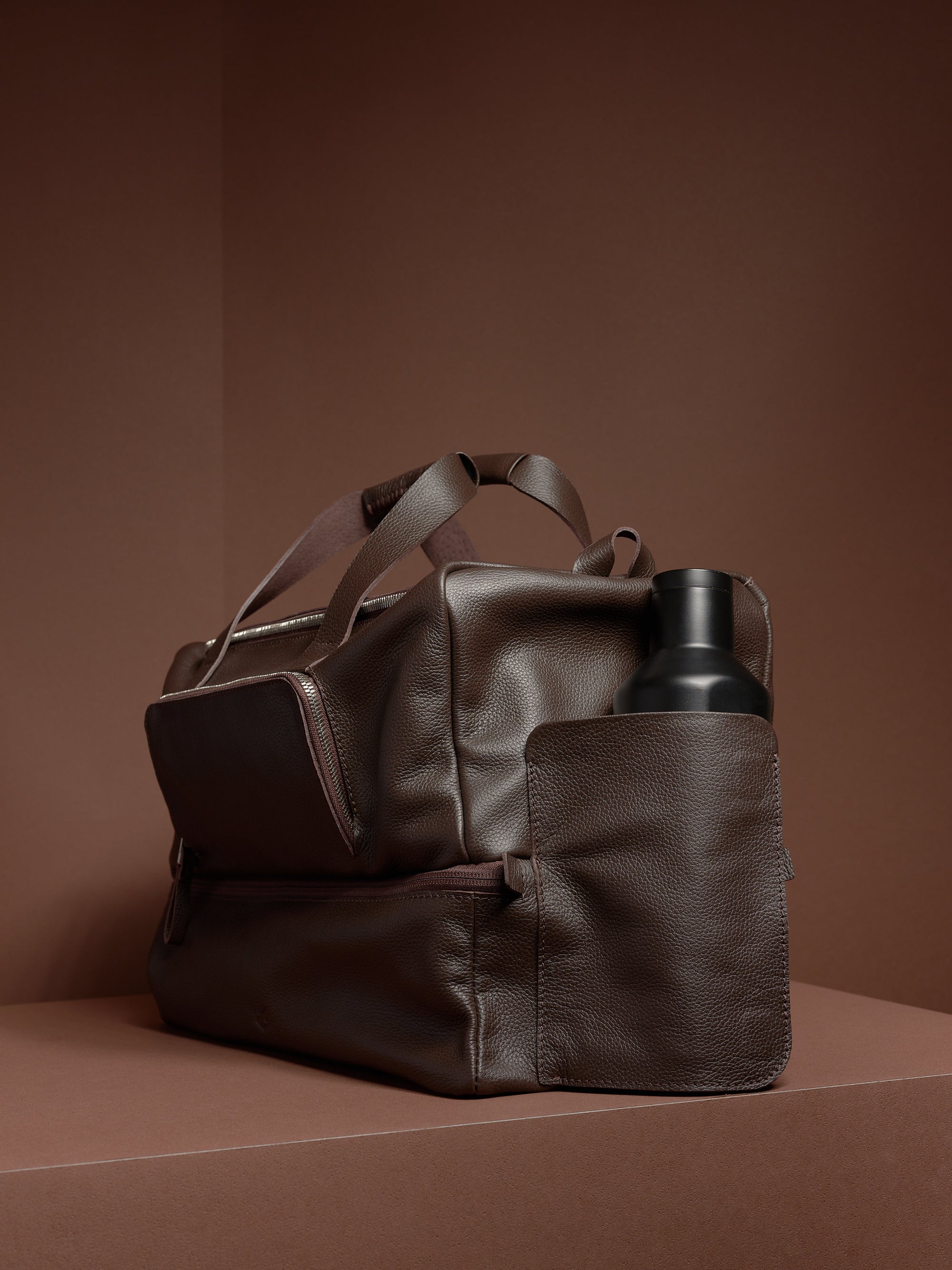 A dark brown leather Ventra Holdall bag on a brown platform with a black water bottle in the side pocket.