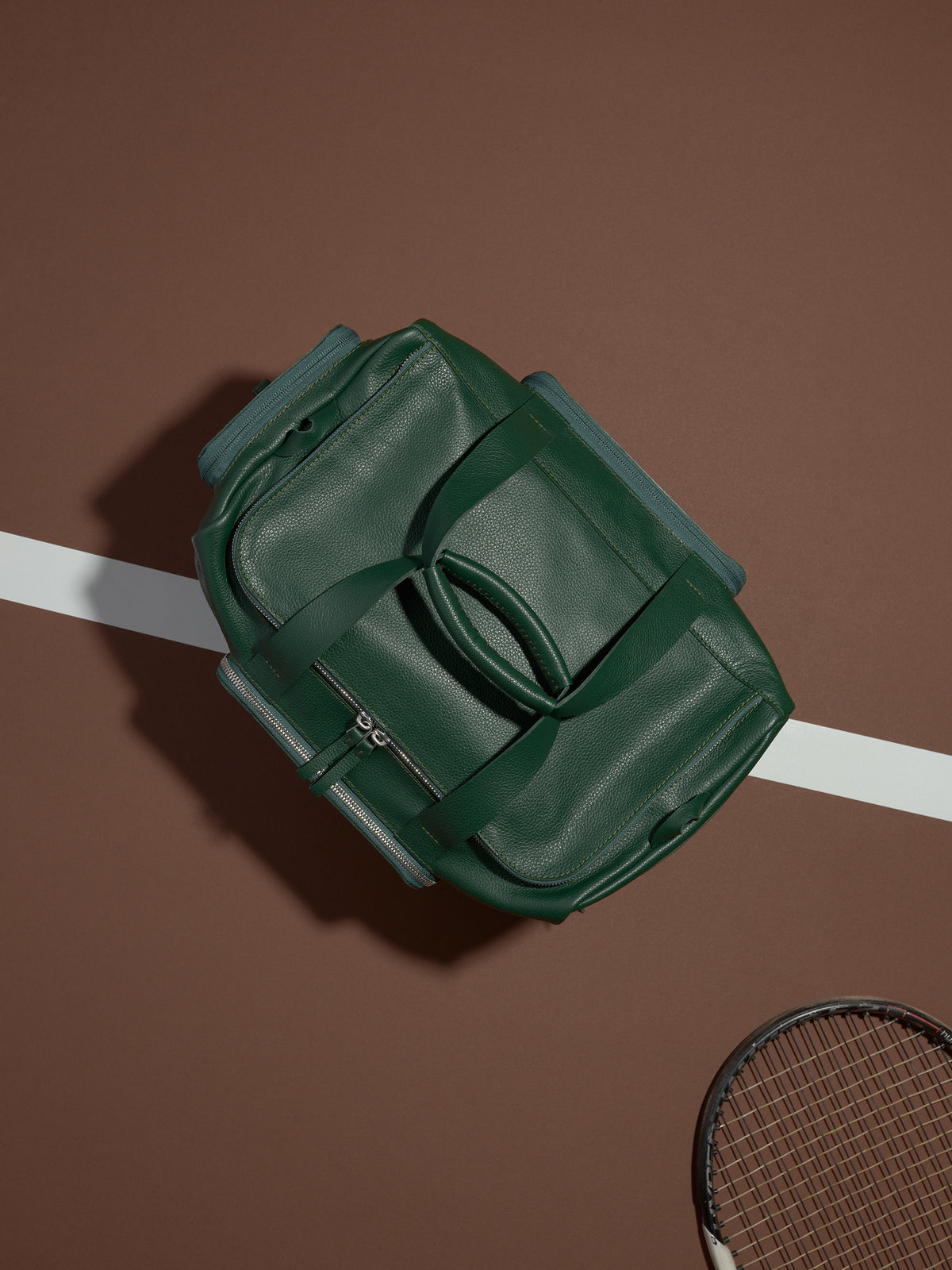 A top-down shot of the green leather Ventra Holdall on a tennis court, positioned as a luxury men's sports bag.