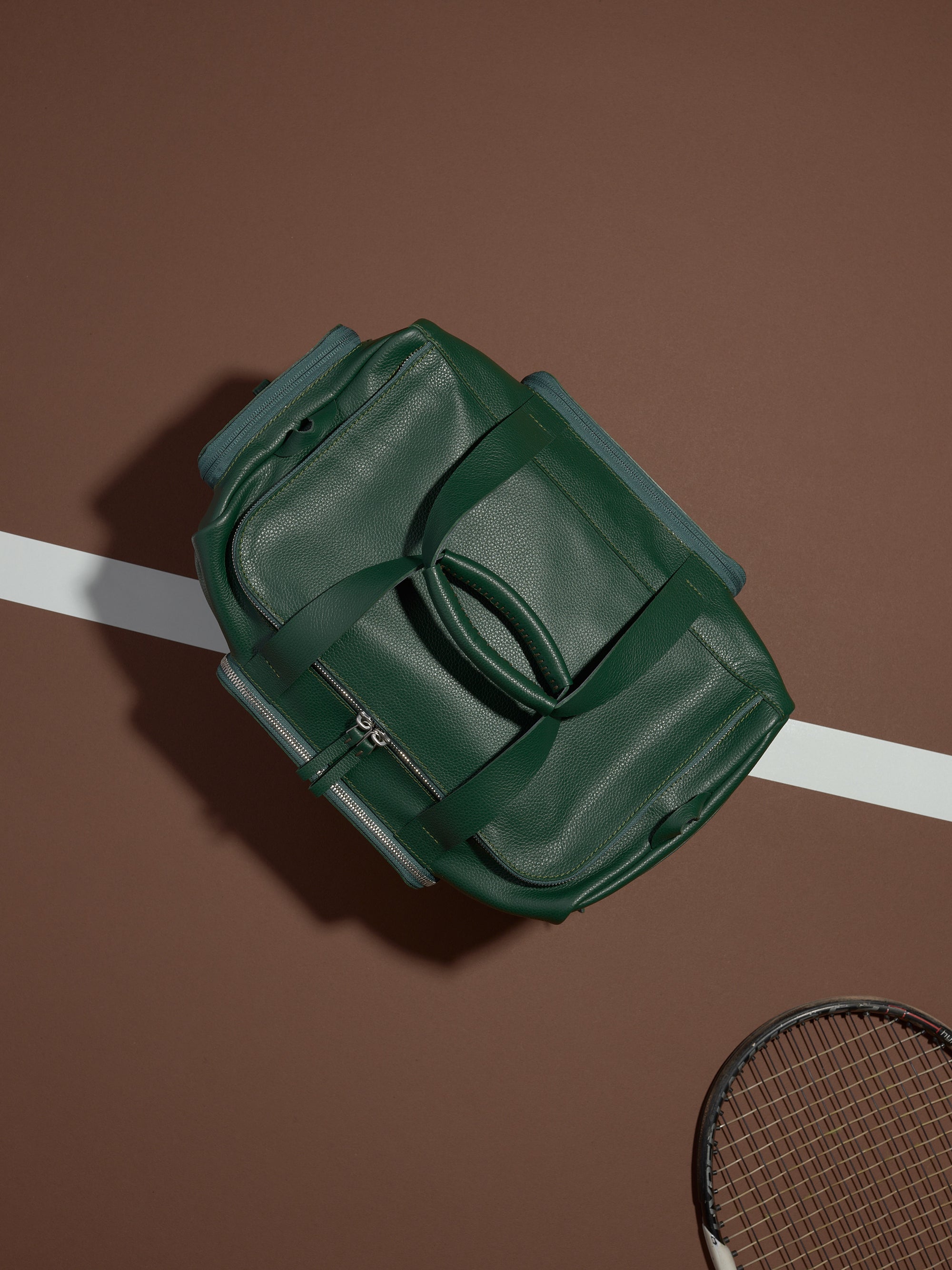A top-down shot of the green leather Ventra Holdall on a tennis court, positioned as a luxury men's sports bag.