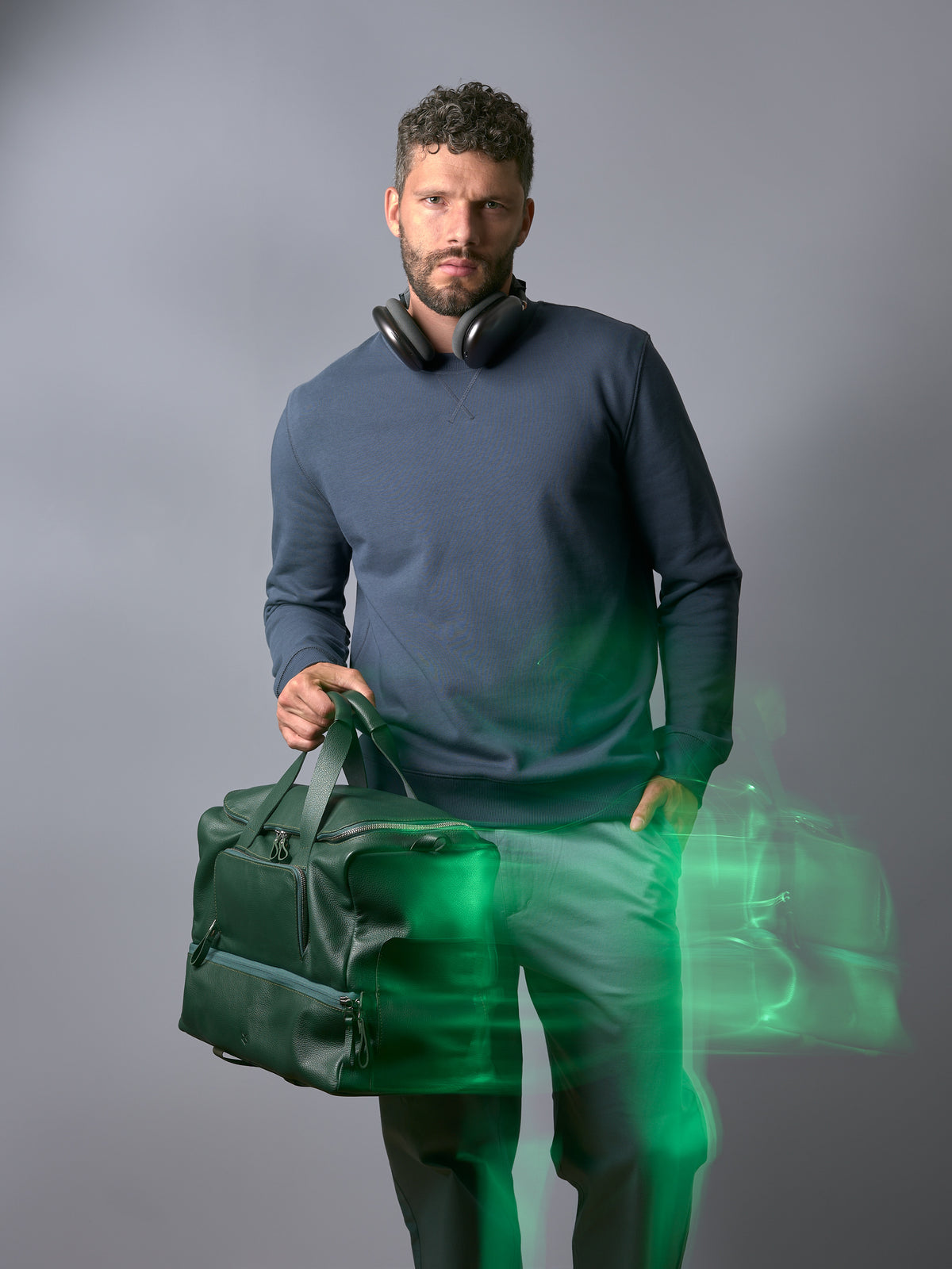 A man with headphones wearing a blue sweatshirt and green pants holding the Capra Leather Ventra Holdall in green.