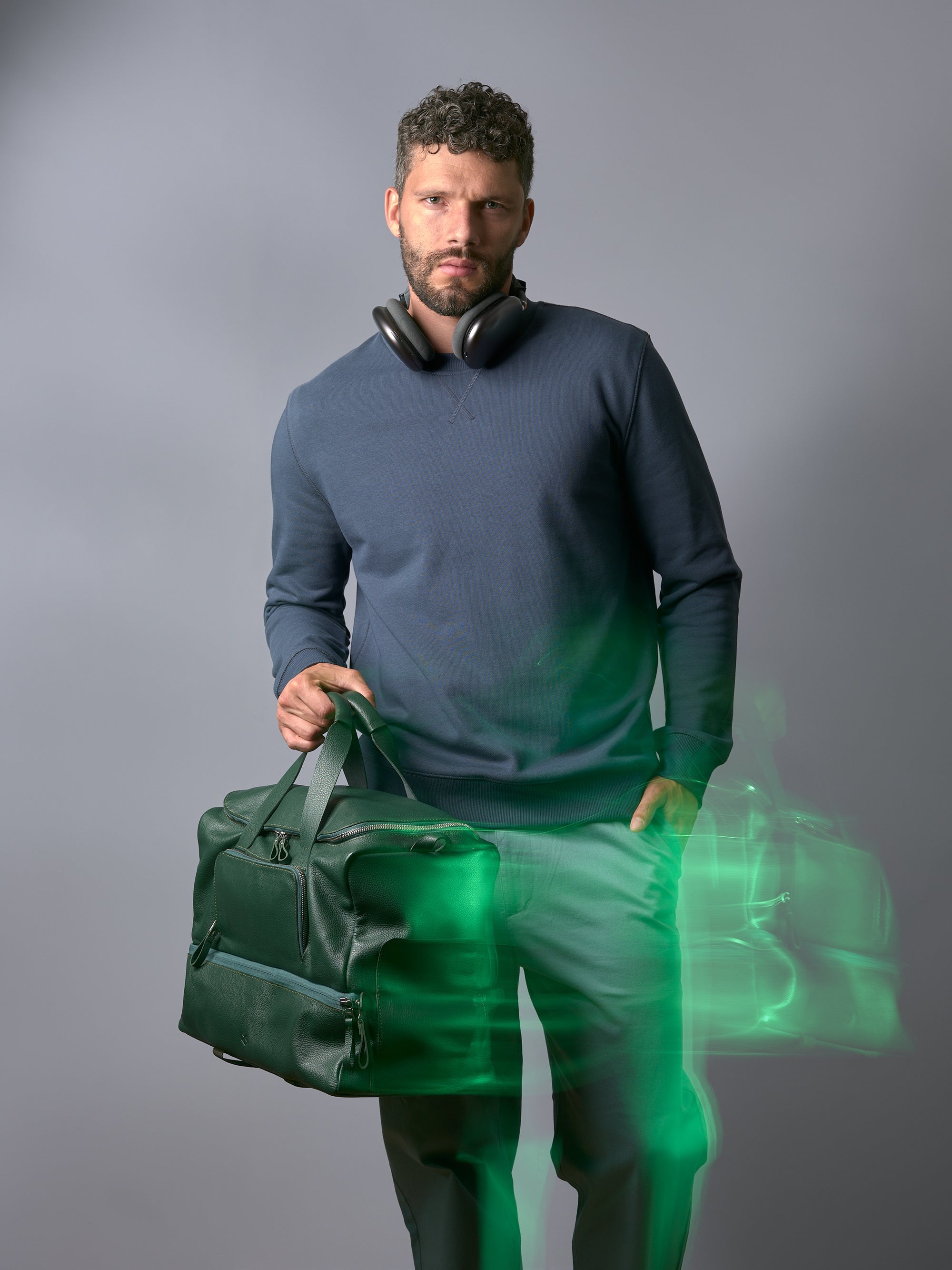 A man with headphones wearing a blue sweatshirt and green pants holding the Capra Leather Ventra Holdall in green.