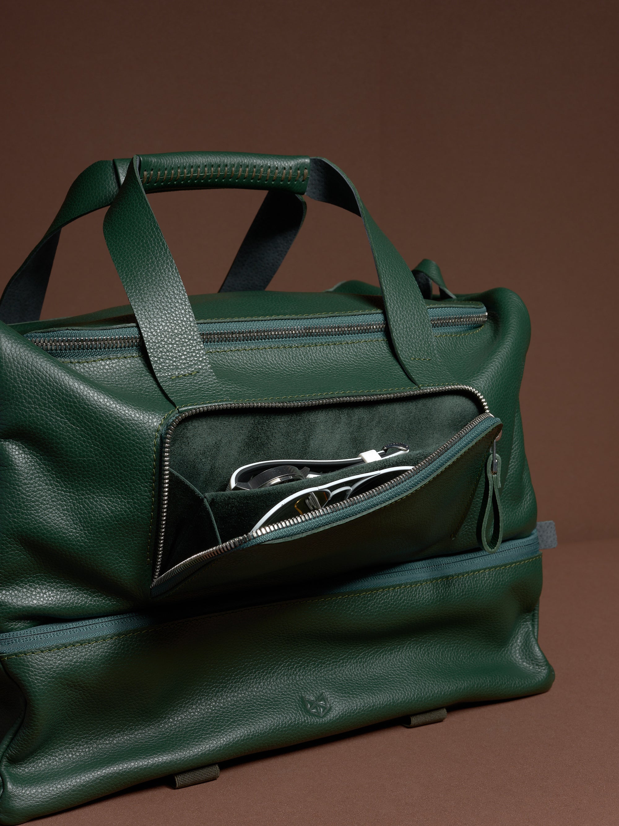 The easy-access front zippered pocket of the green leather Ventra Holdall, open to show wristwatch and glasses inside.