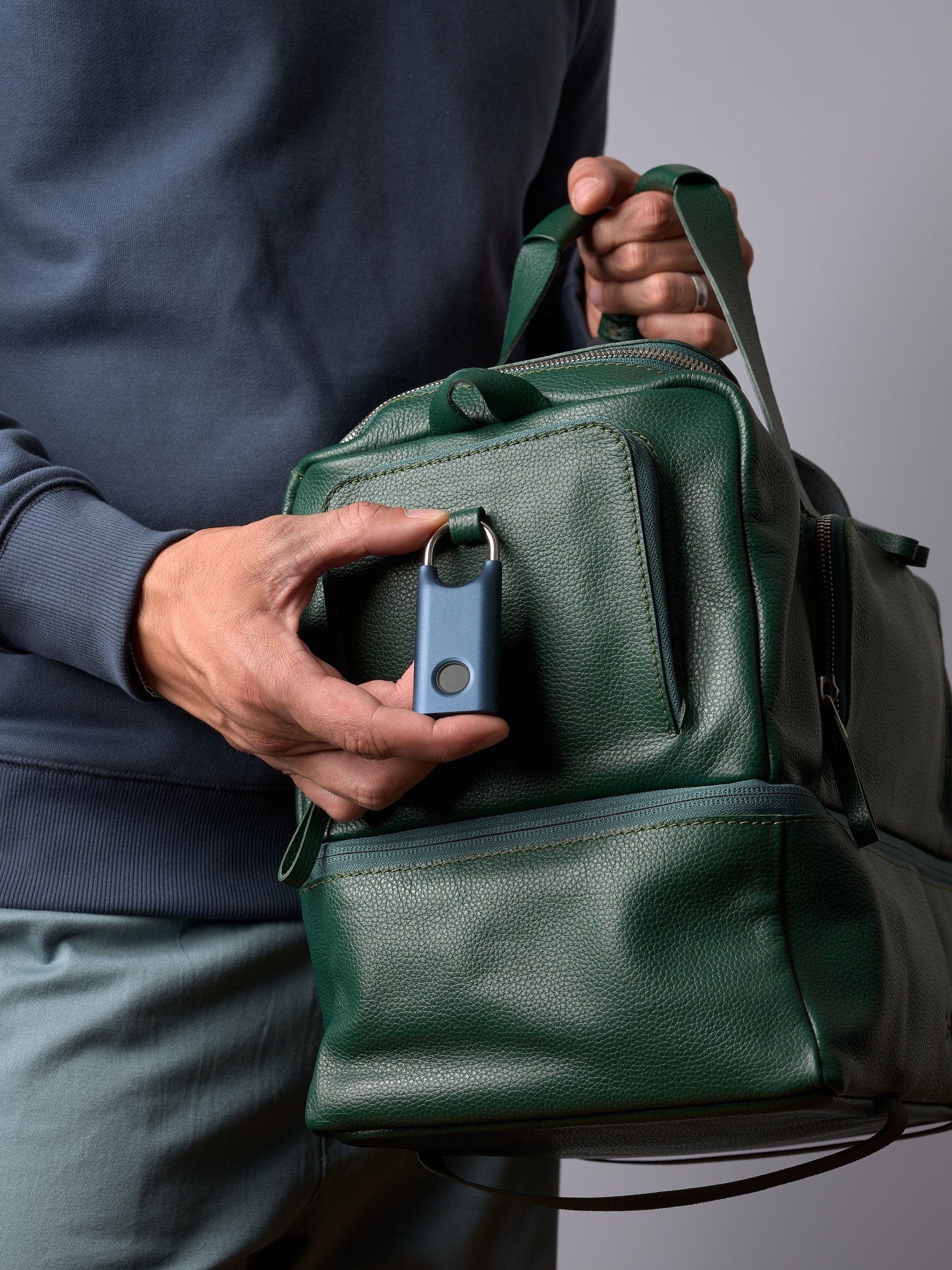 A man's hand holding a blue tech tracker, attaching it to a front loop on the green leather Ventra Holdall bag.