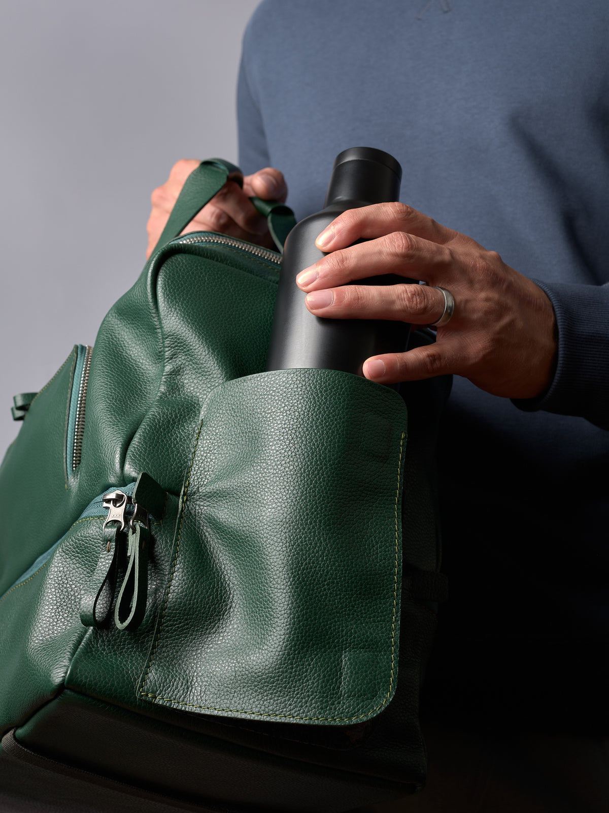 A hand placing a black water bottle into the exterior side pocket of the green leather Ventra Holdall.