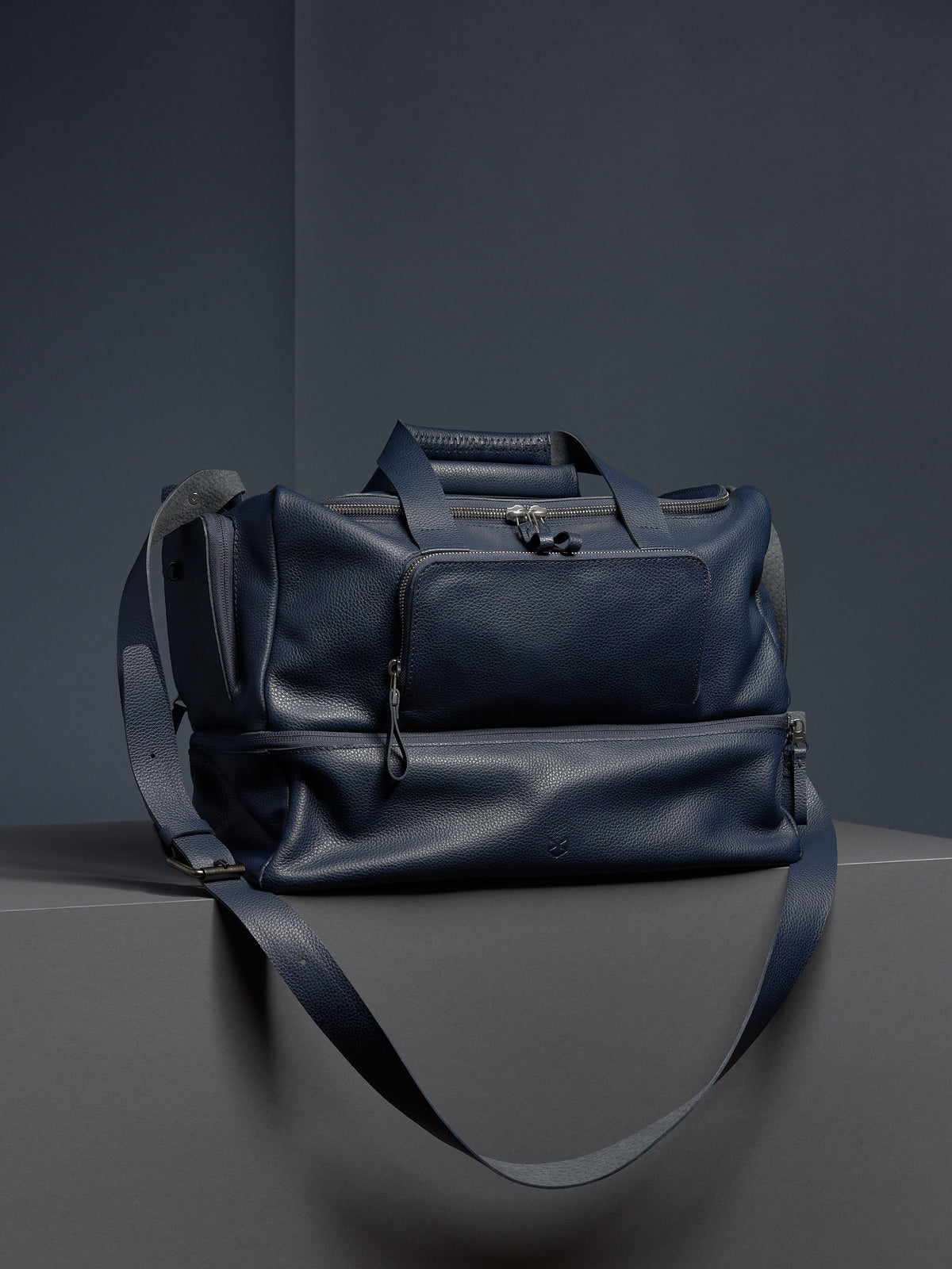 Navy leather Ventra Holdall bag on a grey platform with a strap draped down.