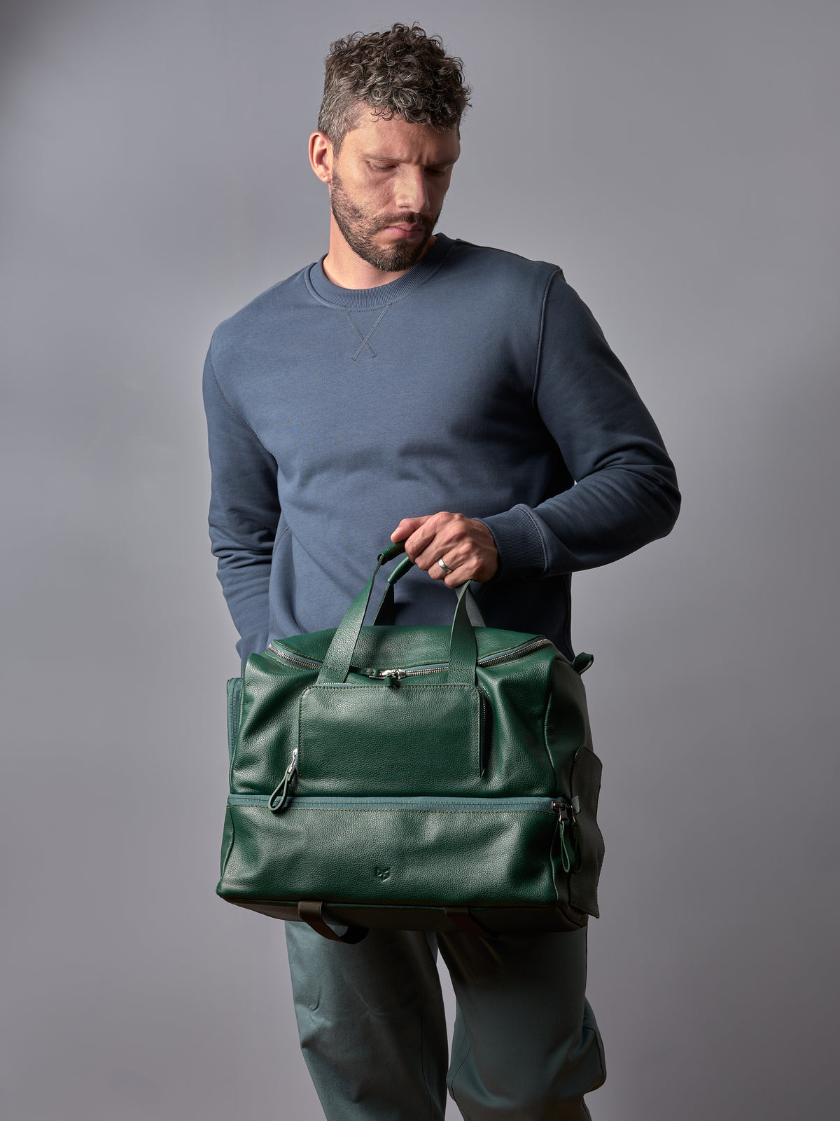 Man in blue sweatshirt and headphones adjusting zipper on a stylish green leather duffle bag from Capra Leather.