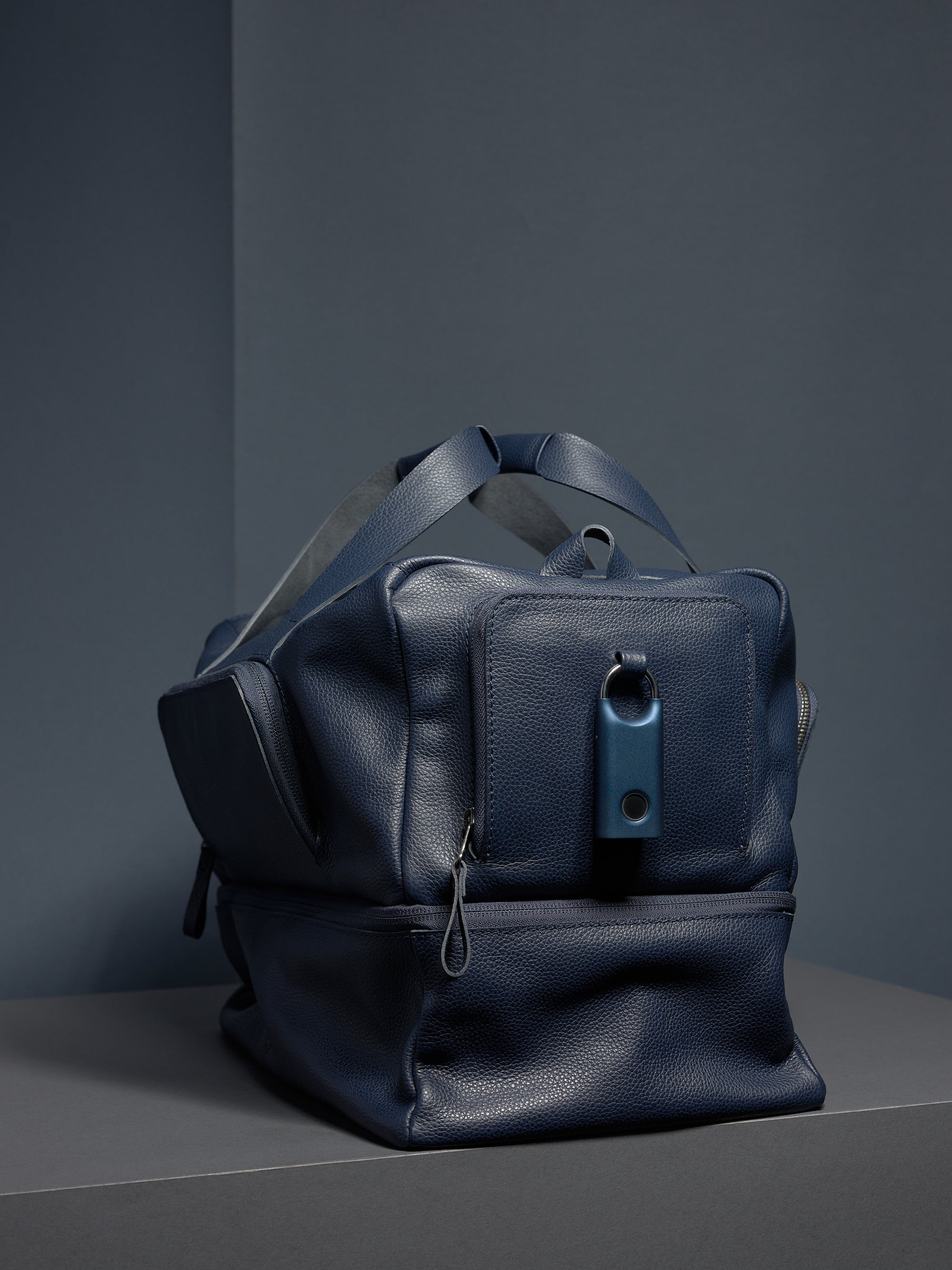 A navy leather Ventra Holdall bag on a grey platform with a blue key tracker attached.