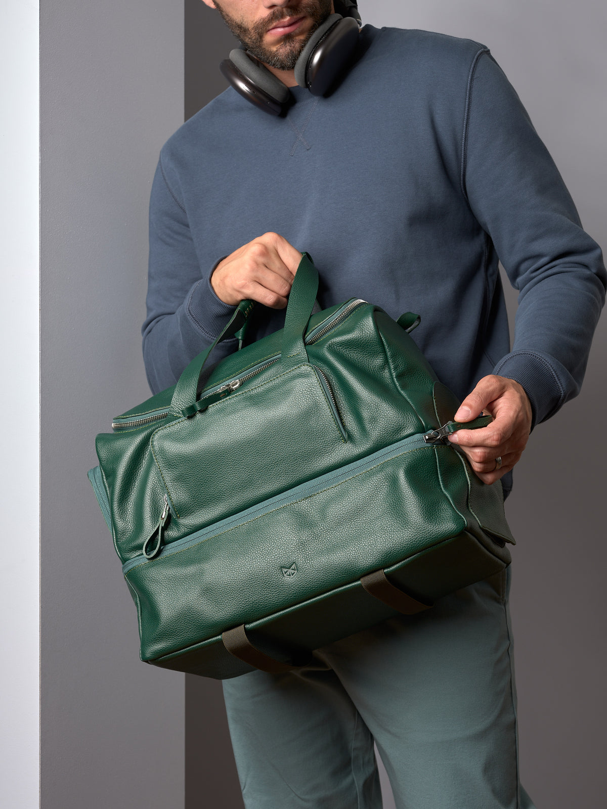 Man with headphones around his neck, looking down at a premium green leather duffle bag from Capra Leather.