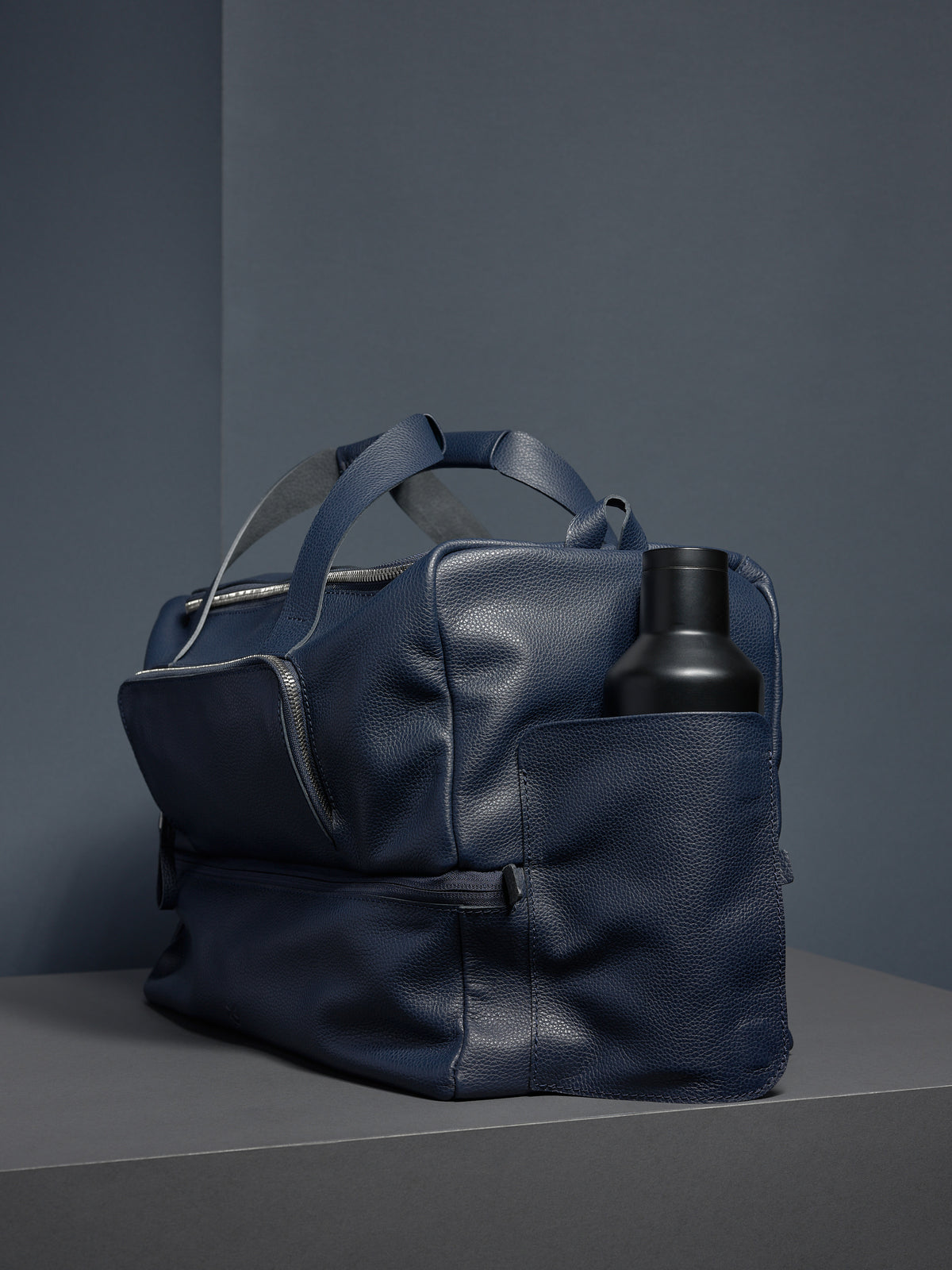 A navy leather Ventra Holdall bag on a grey platform with a black water bottle in the side pocket.