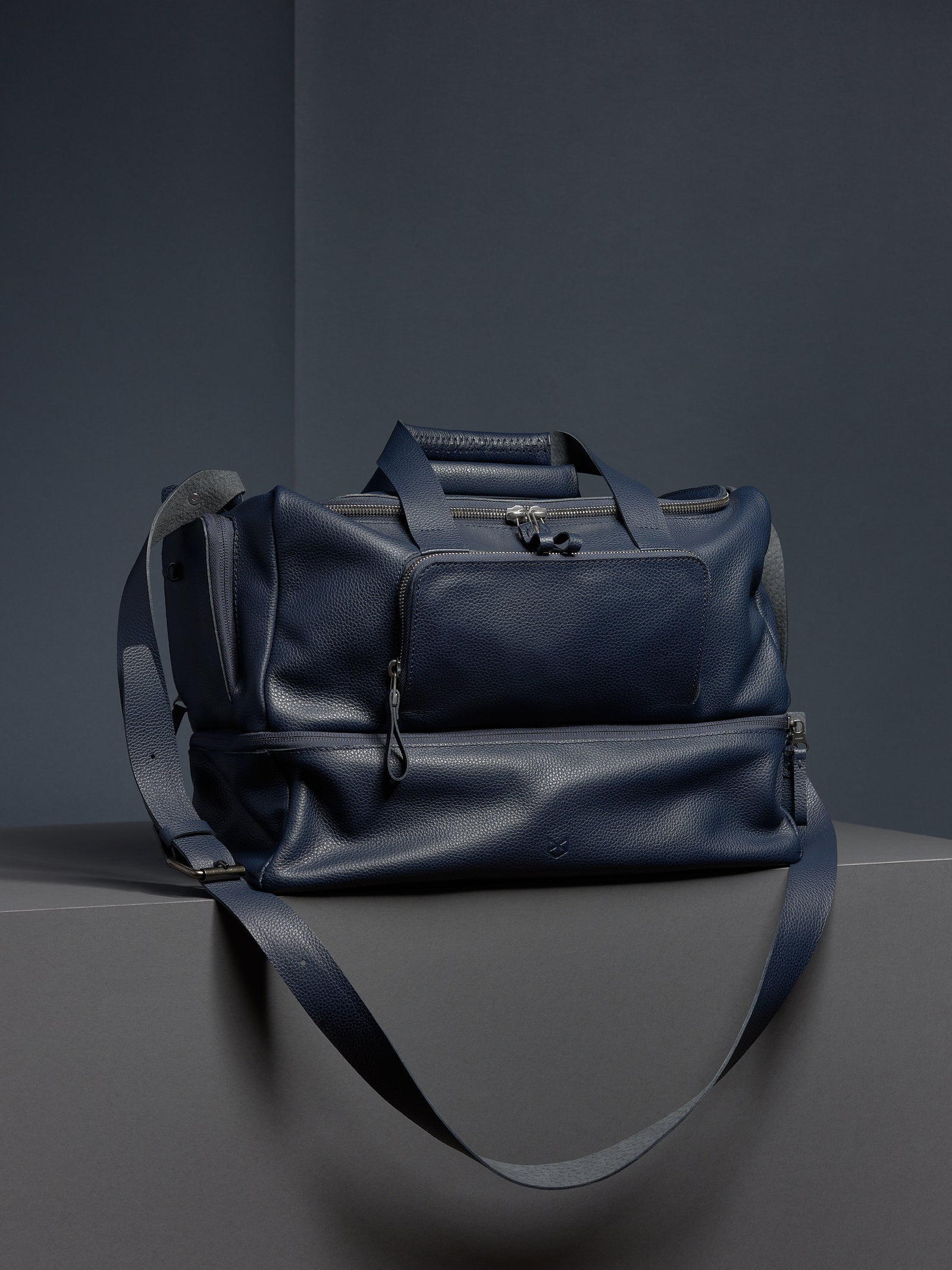 Navy leather Ventra Holdall bag on a grey platform with a strap draped down.