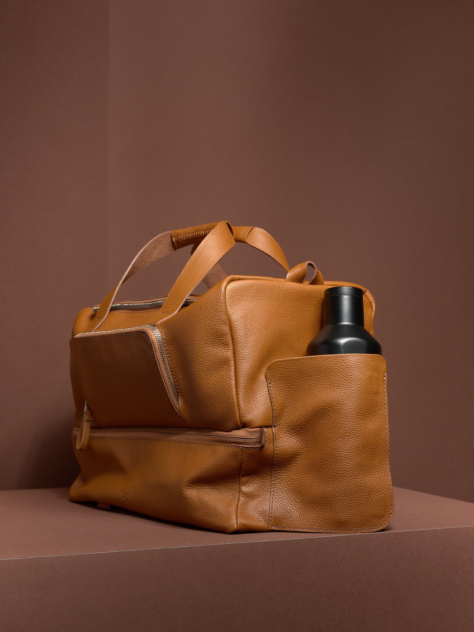 Side view of the tan leather Ventra Holdall duffel bag, showing a black water bottle neatly fitting in the side pocket.