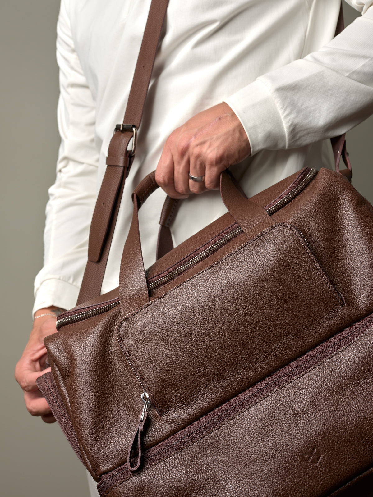 Close-up of a man's hands holding the brown leather Capra Ventra Holdall, highlighting the rich texture and zippered pockets.