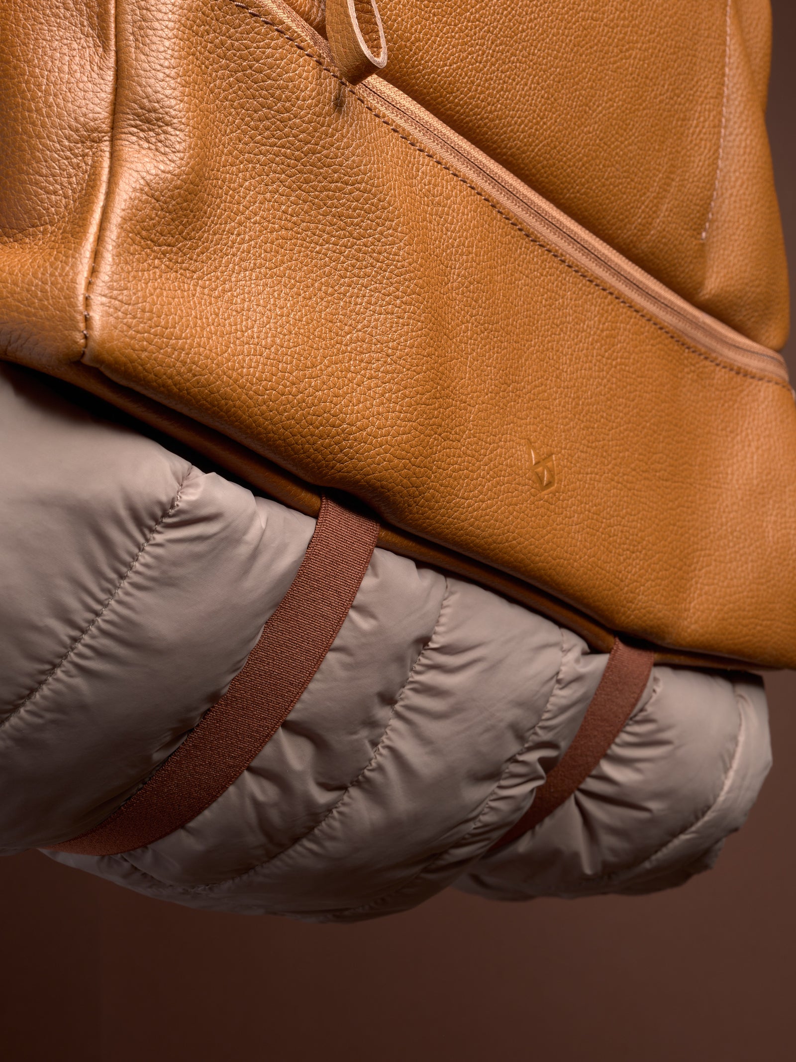 Close-up of the Ventra Holdall's base, where elastic straps securely hold a rolled puffer jacket against the tan leather.