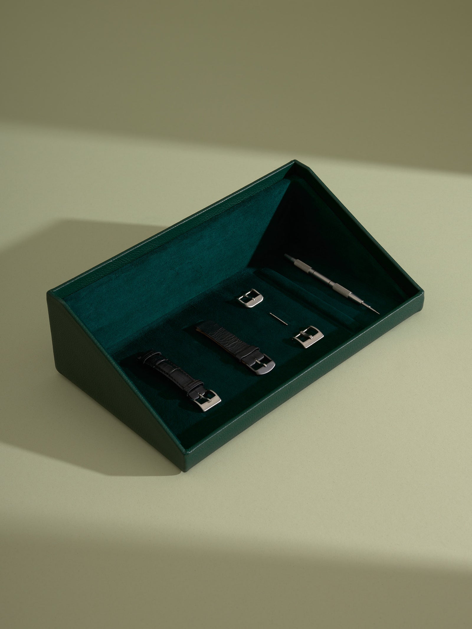 Angled top view of the green leather valet drawer lined with soft green suede, holding watch straps, buckles, and a strap changing tool.
