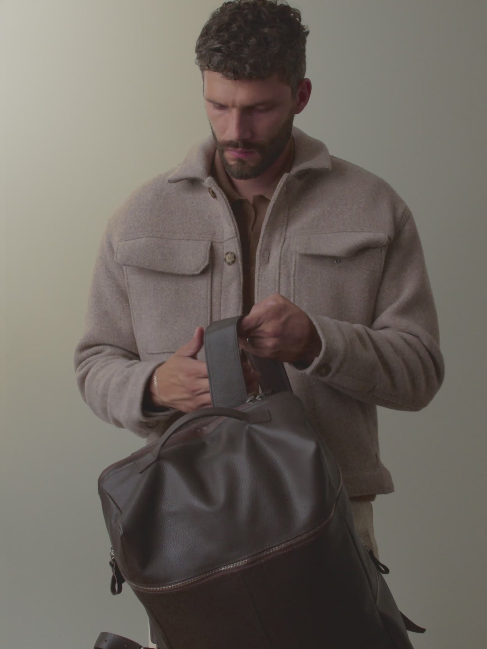 A stylish man wearing a beige jacket demonstrates the features of the Capra Leather Banteng full-grain leather backpack in brown, including zipping compartments, stowing a phone and water bottle, and clipping sunglasses to the shoulder strap before wearing the backpack.