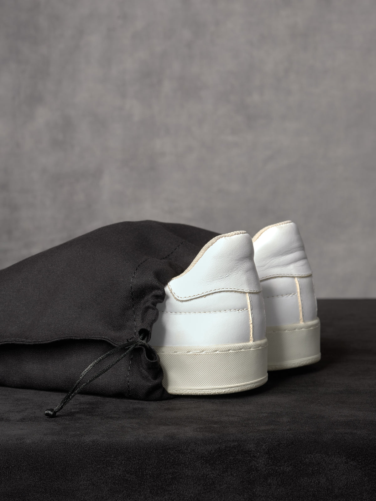 A pair of white leather sneakers partially tucked into a black canvas drawstring shoe bag on a dark, textured surface.