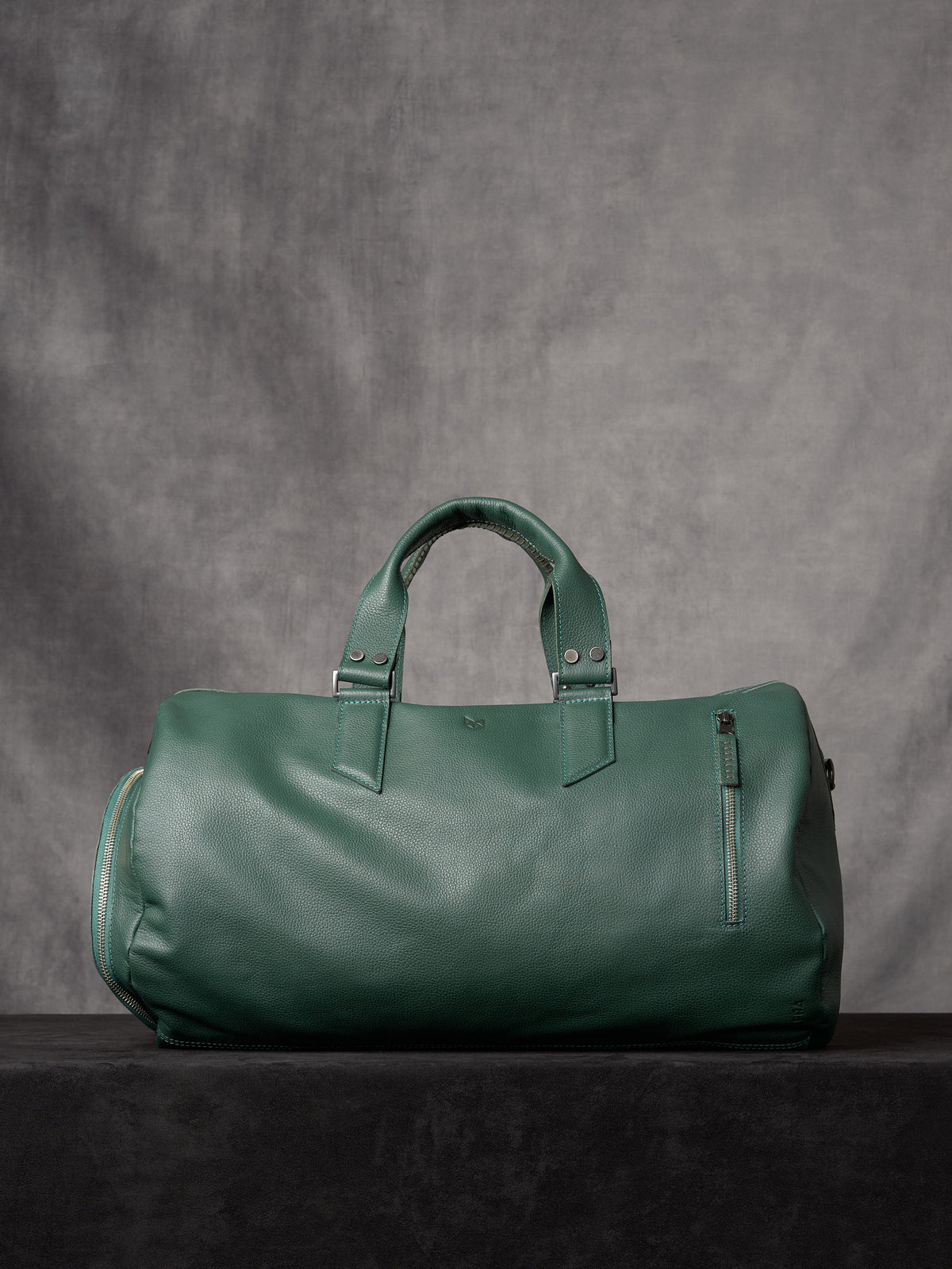 Front view of a forest green leather duffle bag with a vertical zipper pocket on a dark pedestal.