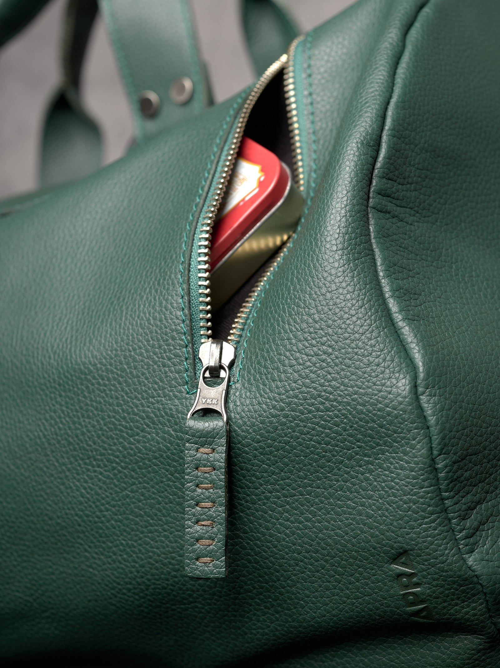 Front view of a forest green leather duffle bag with a vertical zipper pocket on a dark pedestal.