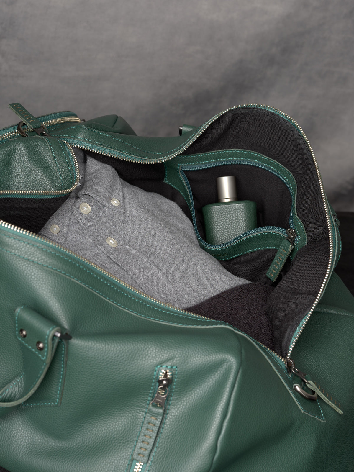 Interior view of the green leather duffle bag packed with a grey shirt and a leather-wrapped fragrance bottle.