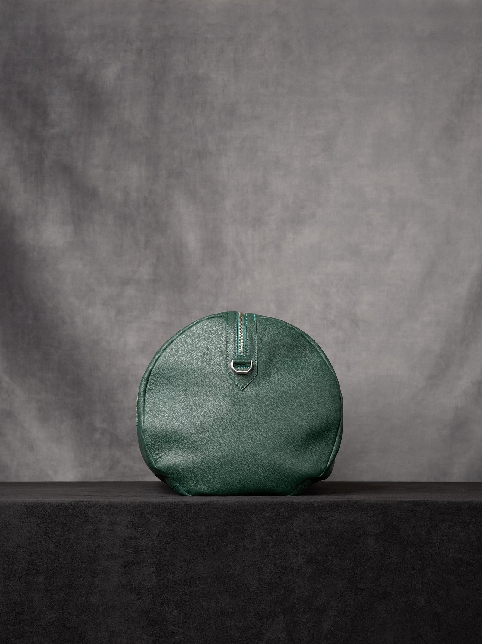 Side view of the forest green leather duffle bag showing its circular profile and a silver D-ring attachment.