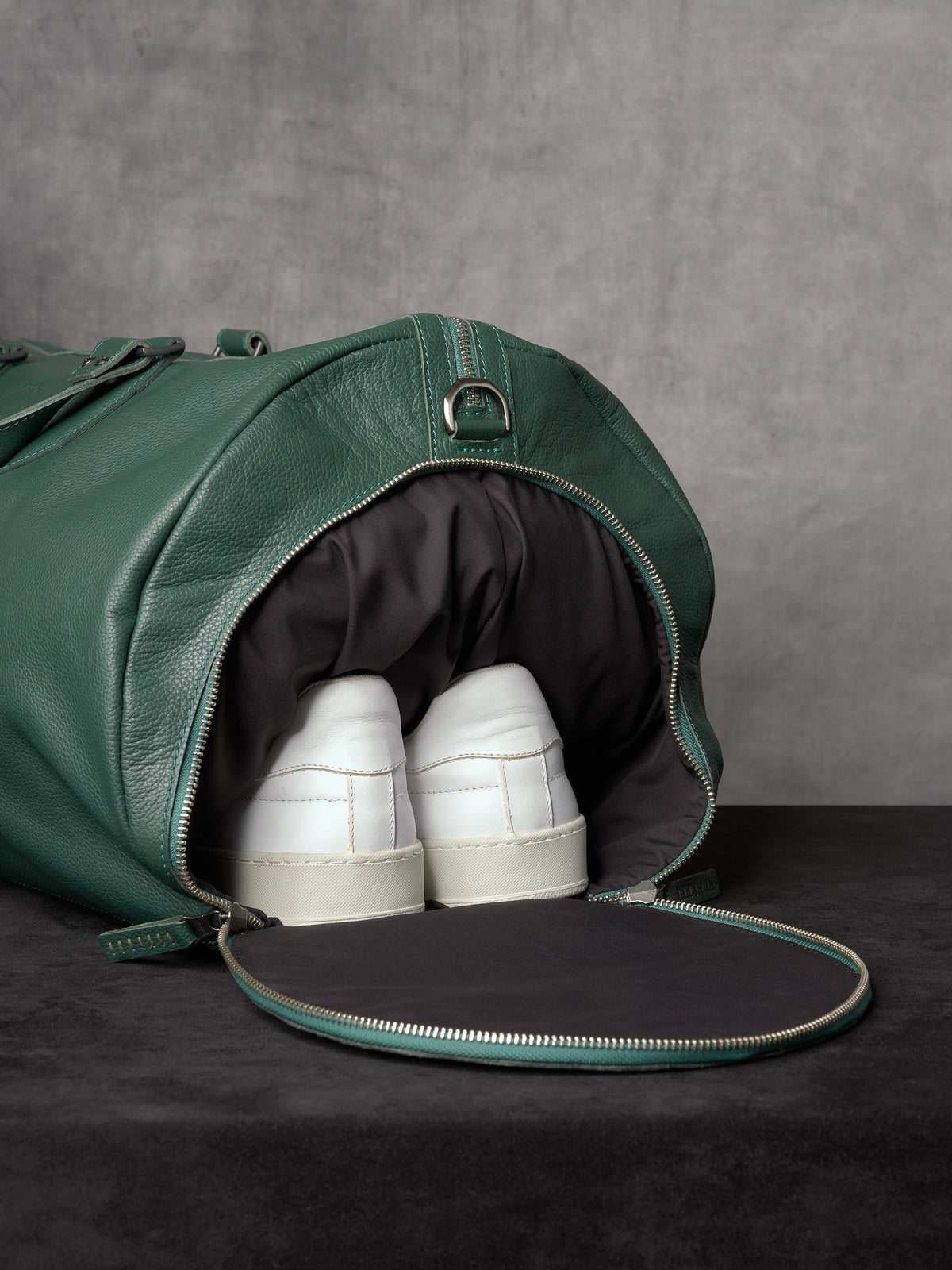 A side compartment of the green leather duffle bag open to reveal a pair of white sneakers stored in a dedicated shoe pocket.