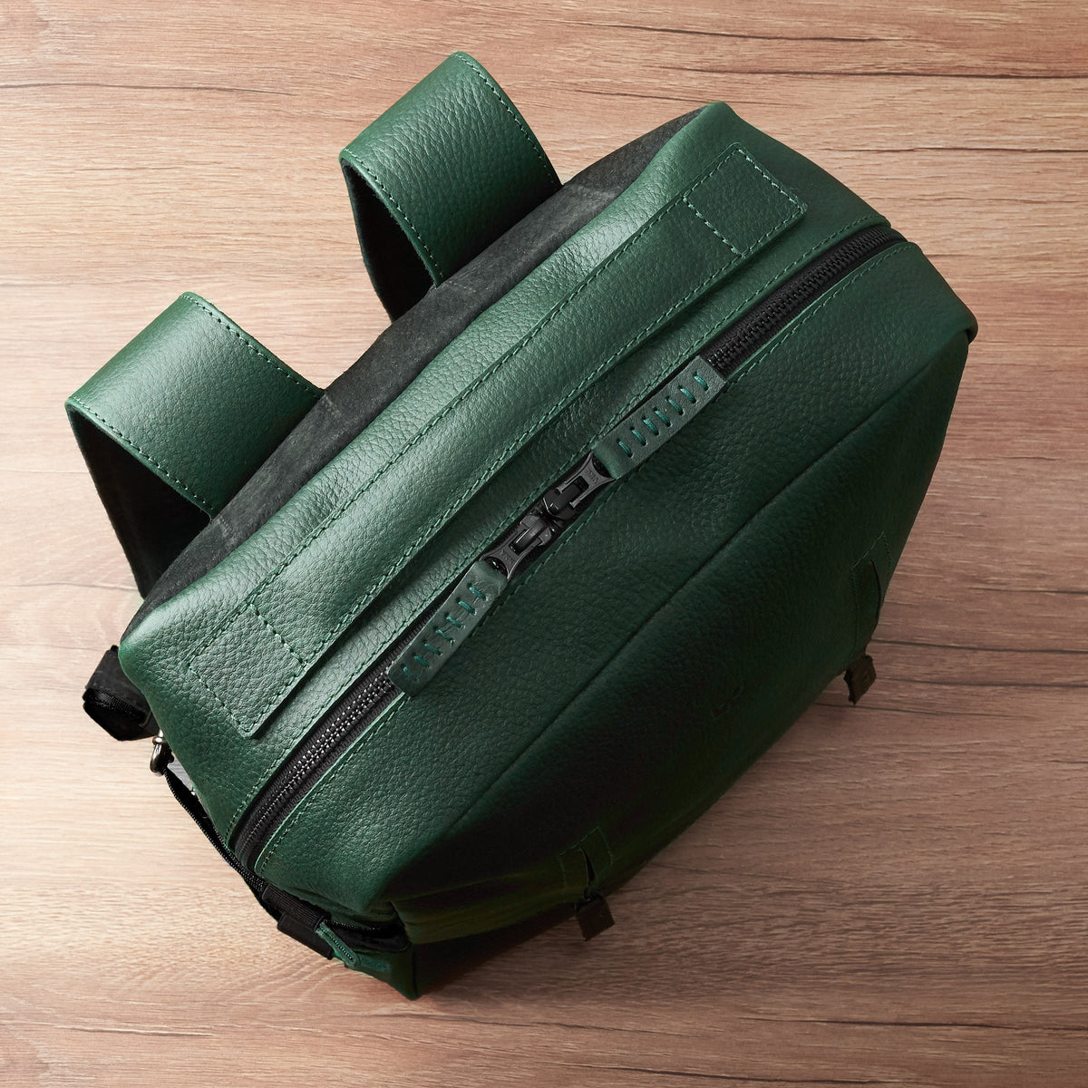 An overhead, angled view of a rectangular green, pebble-grain leather backpack lying on a wooden surface, showing the main zipper closure and the leather-covered top handle.