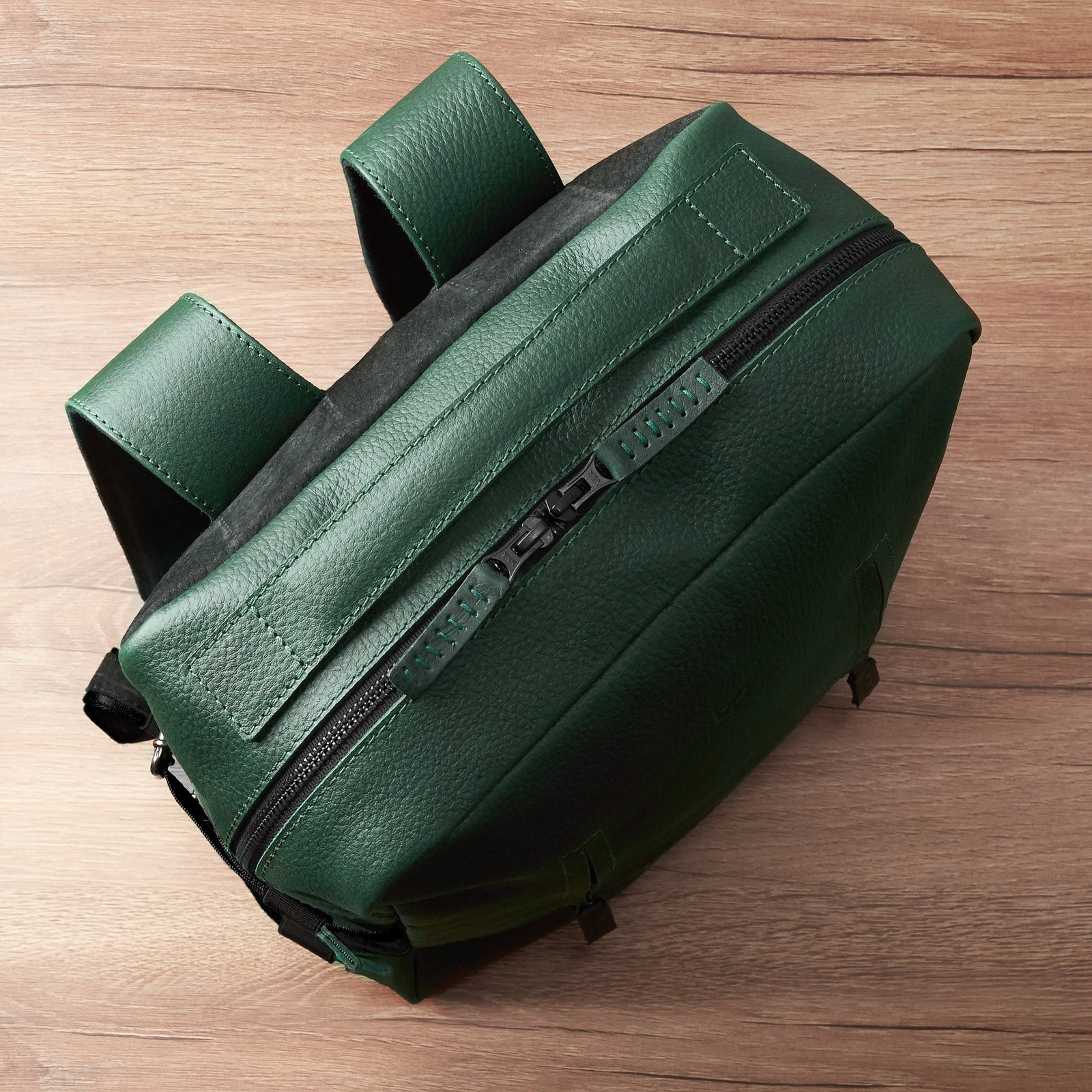 An overhead, angled view of a rectangular green, pebble-grain leather backpack lying on a wooden surface, showing the main zipper closure and the leather-covered top handle.