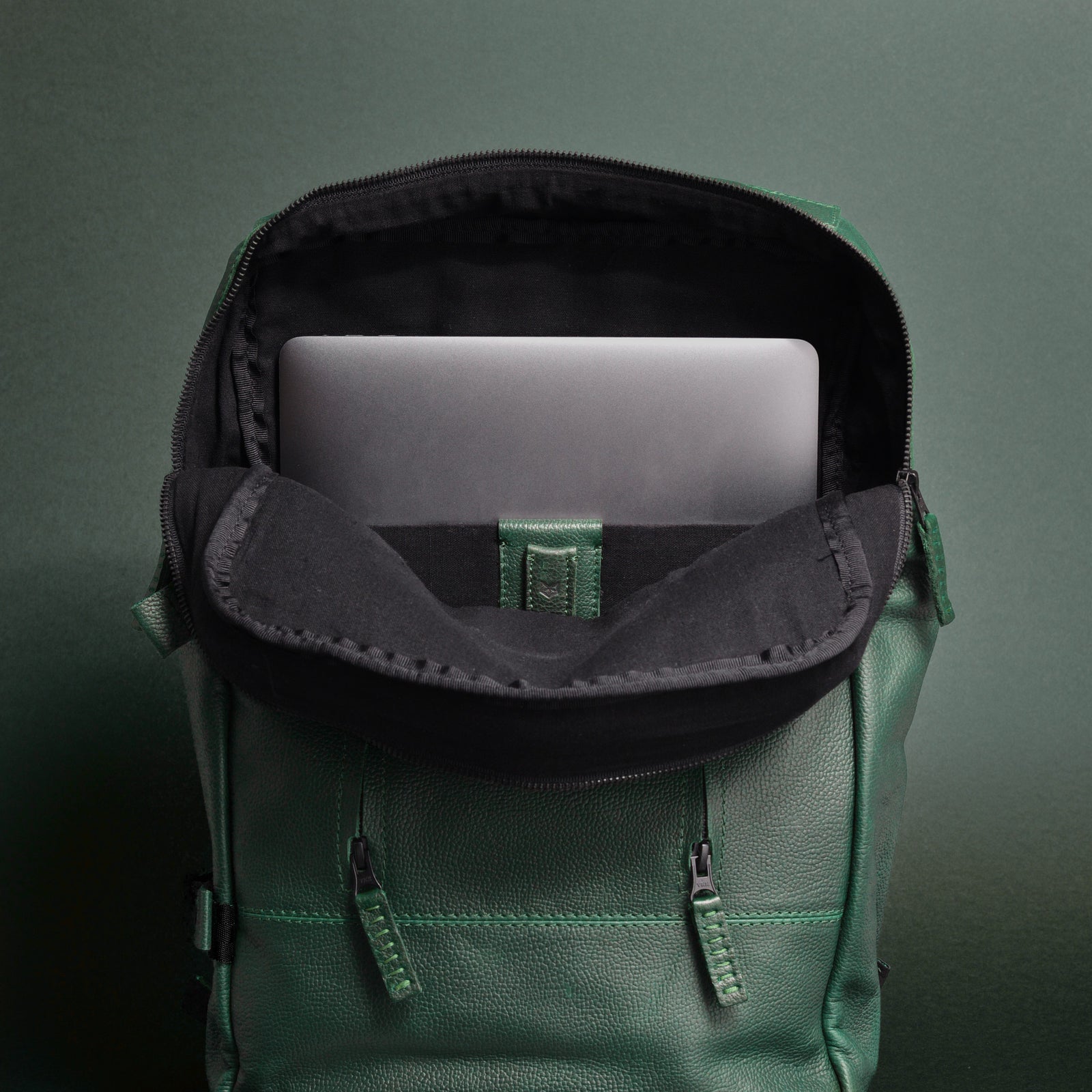 The interior of an open, rectangular green leather backpack against a dark green background. A dark gray laptop is visible in the black padded sleeve.
