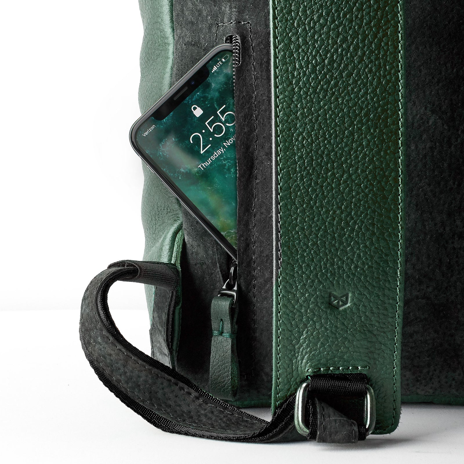 A close-up of a vertical zipper pocket on the side of a rectangular green leather backpack, with a black smartphone partially inserted, showing the screen time.
