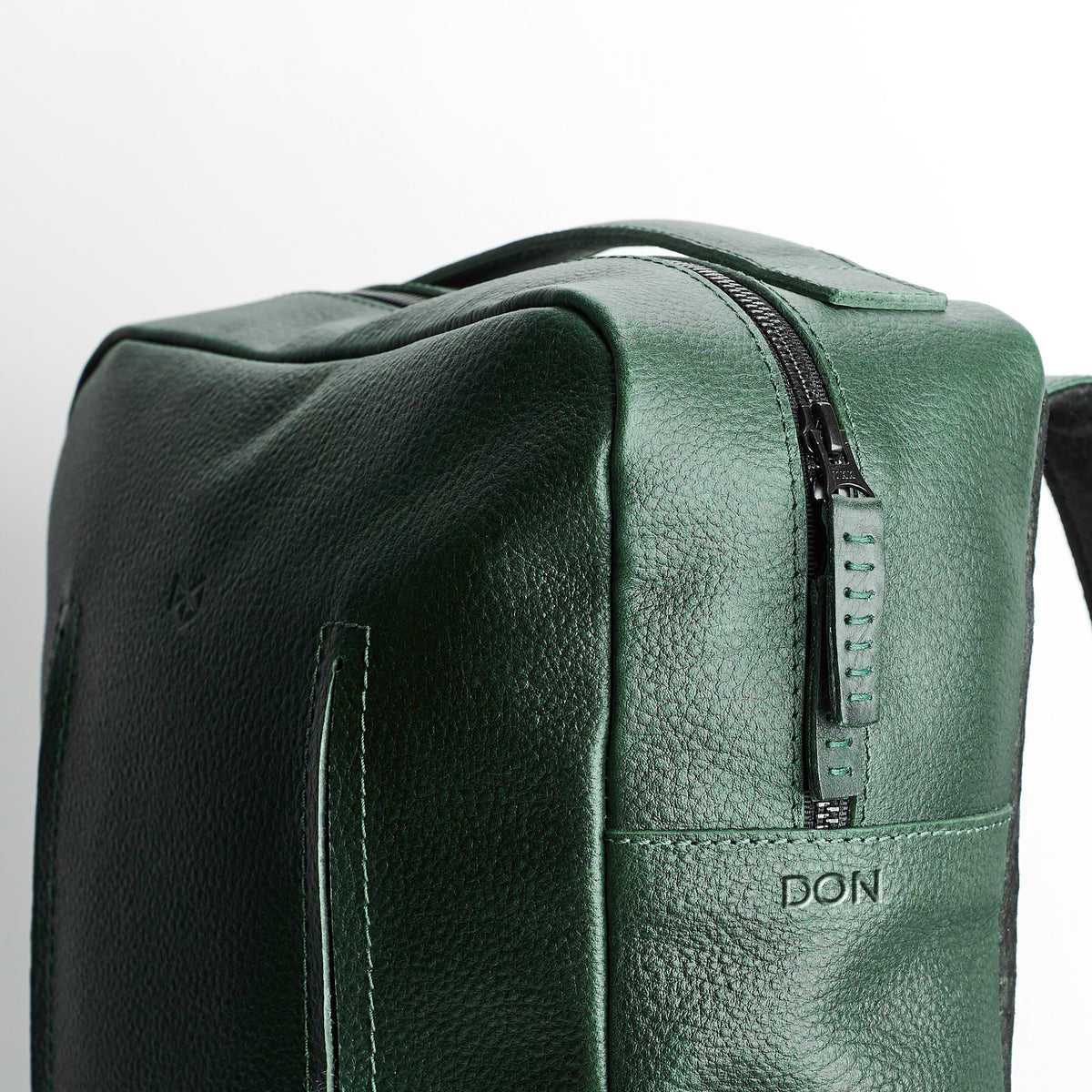 A three-quarter view close-up of the rectangular green leather bag, highlighting the top zipper and the embossed word "DON" near the bottom front panel.