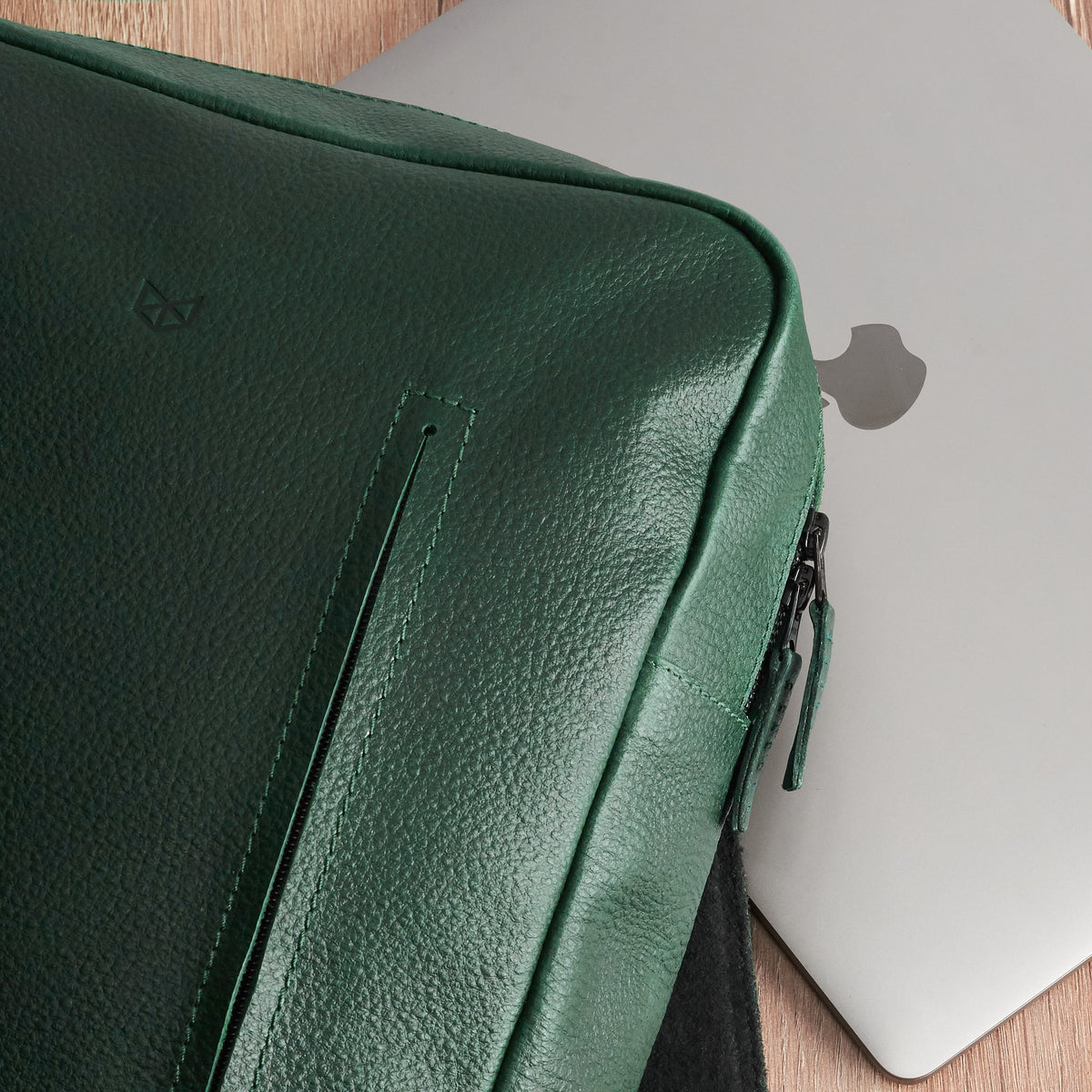 A close-up of the green leather backpack resting on a wooden surface next to a silver laptop, highlighting the bag's pebbled texture and a front seam.