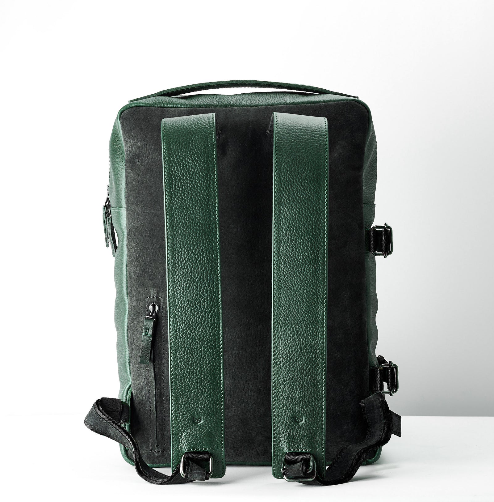 The back view of a rectangular green leather backpack, showing two wide green leather shoulder straps, a black suede-like back panel, and black compression/side straps.
