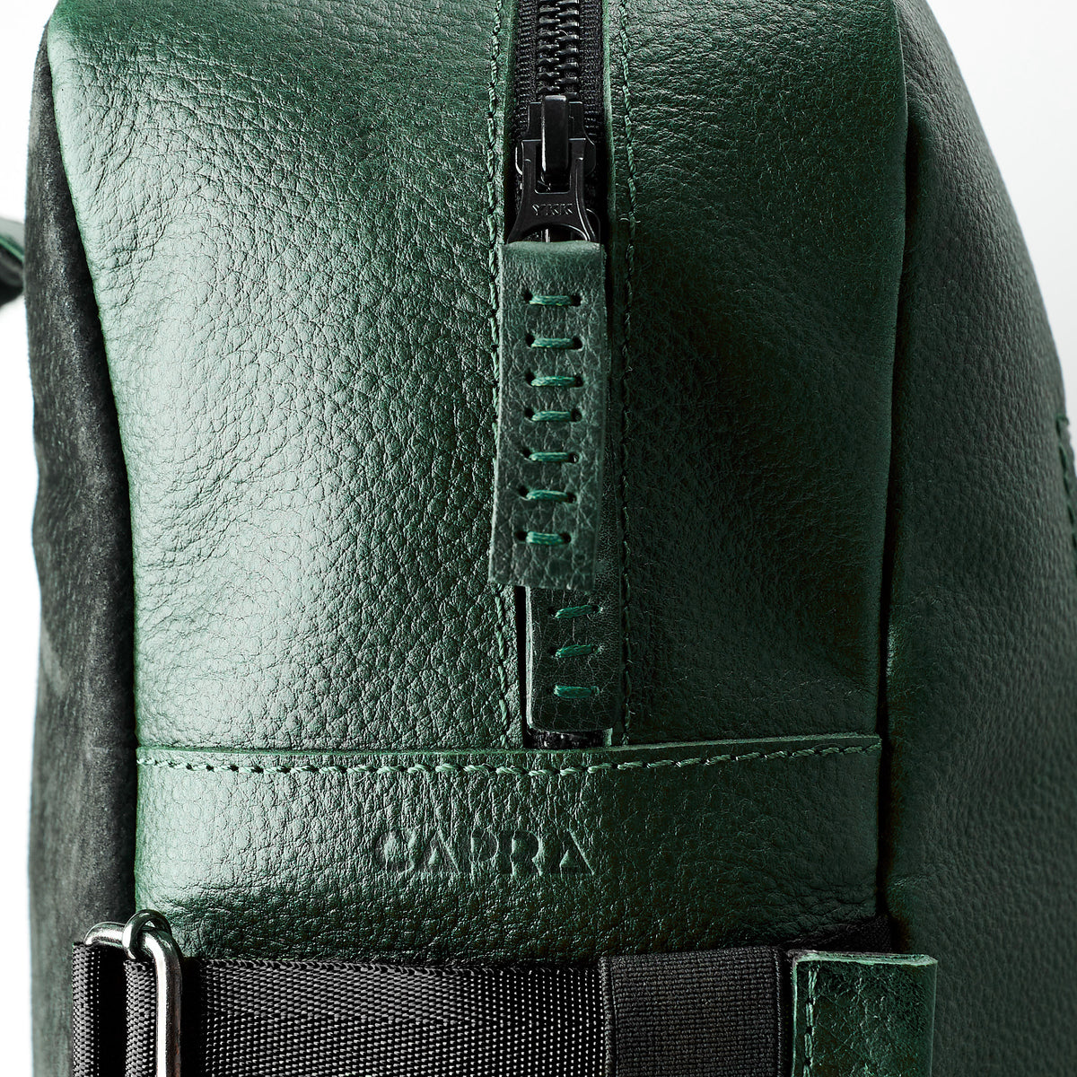 An extreme close-up detail of a black YKK zipper and the green leather pull tab on the bag, with the word "CAPRA" embossed into the leather just below the zipper line.