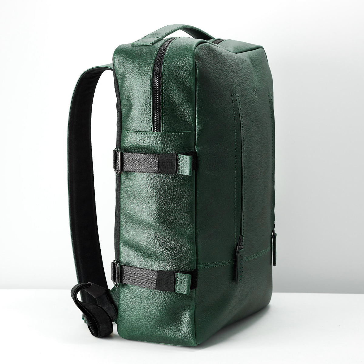 A side view of the rectangular green leather backpack, showing the adjustable shoulder strap and the black compression straps with buckles on the side.