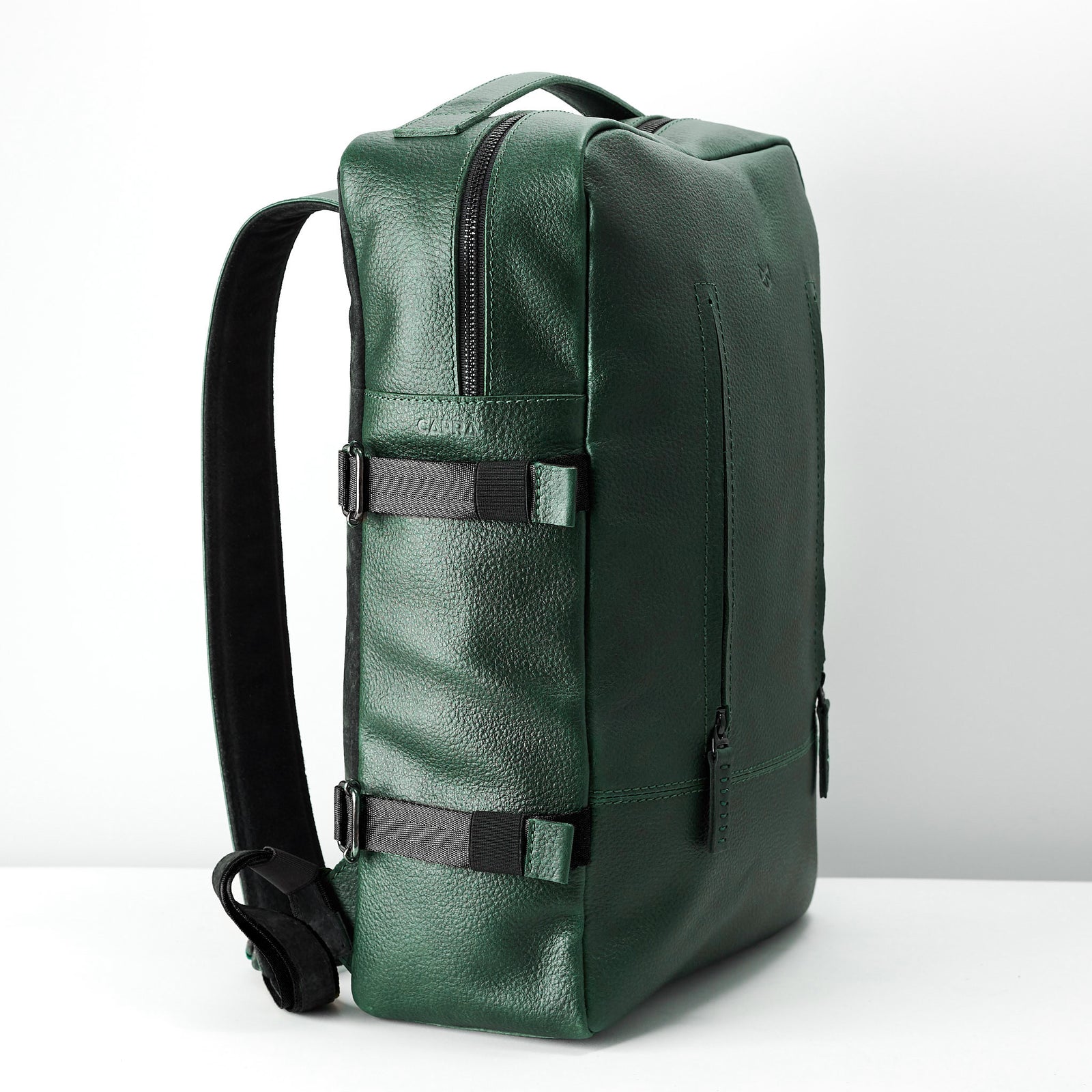A side view of the rectangular green leather backpack, showing the adjustable shoulder strap and the black compression straps with buckles on the side.