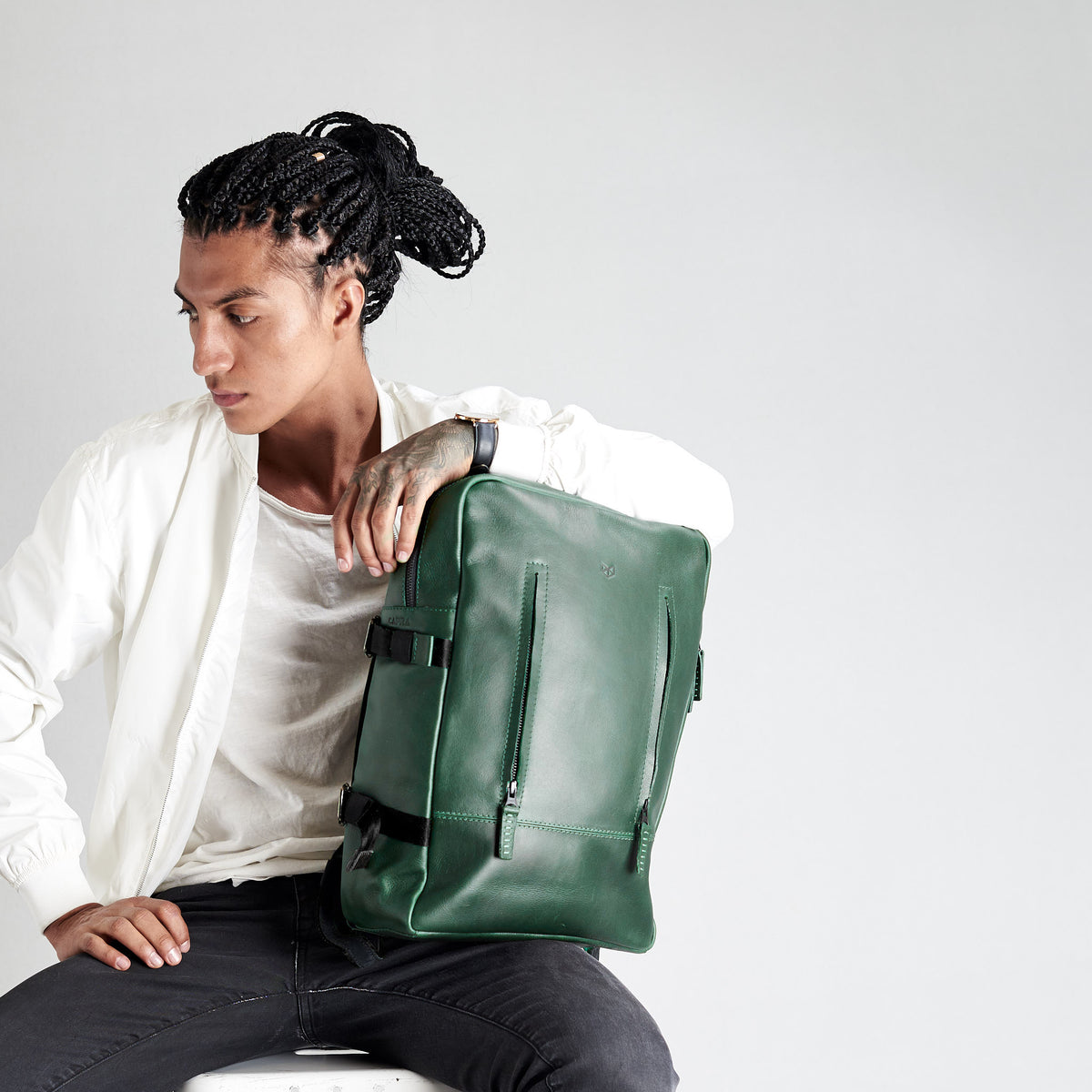 A young man with black braids, wearing a white jacket, sitting and leaning on the rectangular green, pebble-grain leather backpack, which is standing upright.
